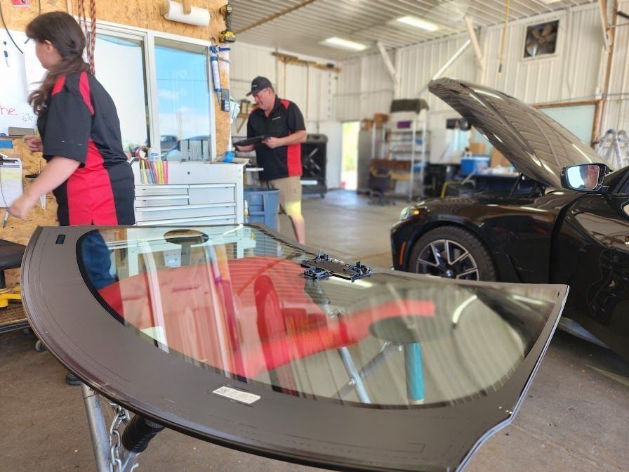 An auto repair shop with a windshield in the foreground and two technicians working on a car with its hood open.