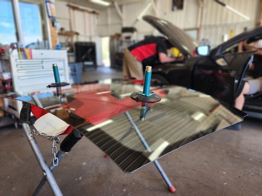 A car windshield with two suction cup handles sits on a stand in a mechanic's garage with a car being repaired nearby.