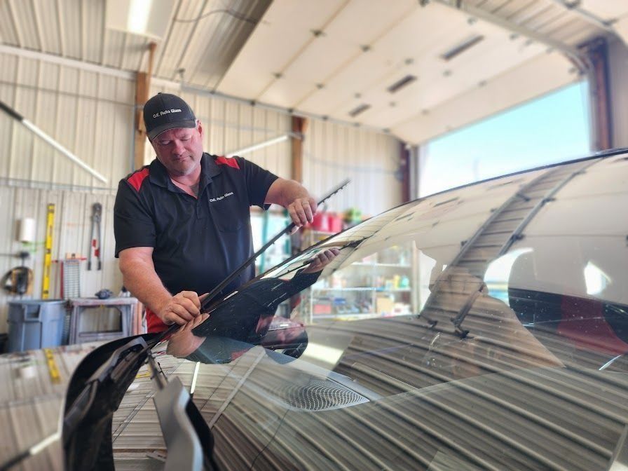 A technician in a black polo shirt installs new windshield wiper blades on a car inside an auto repair shop.