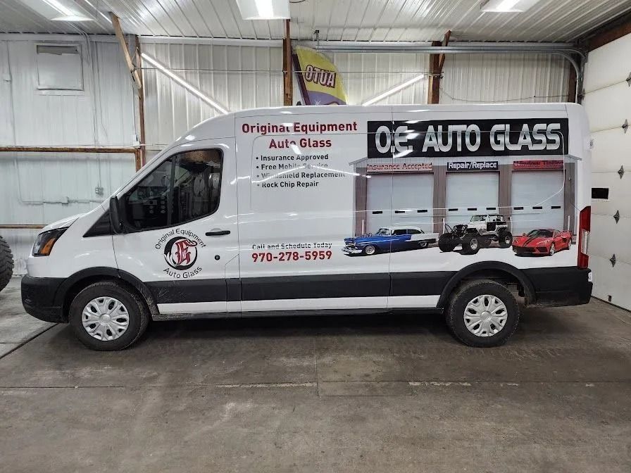 A white O.E. Auto Glass work van parked inside a workshop, featuring business branding and car graphics on its side.