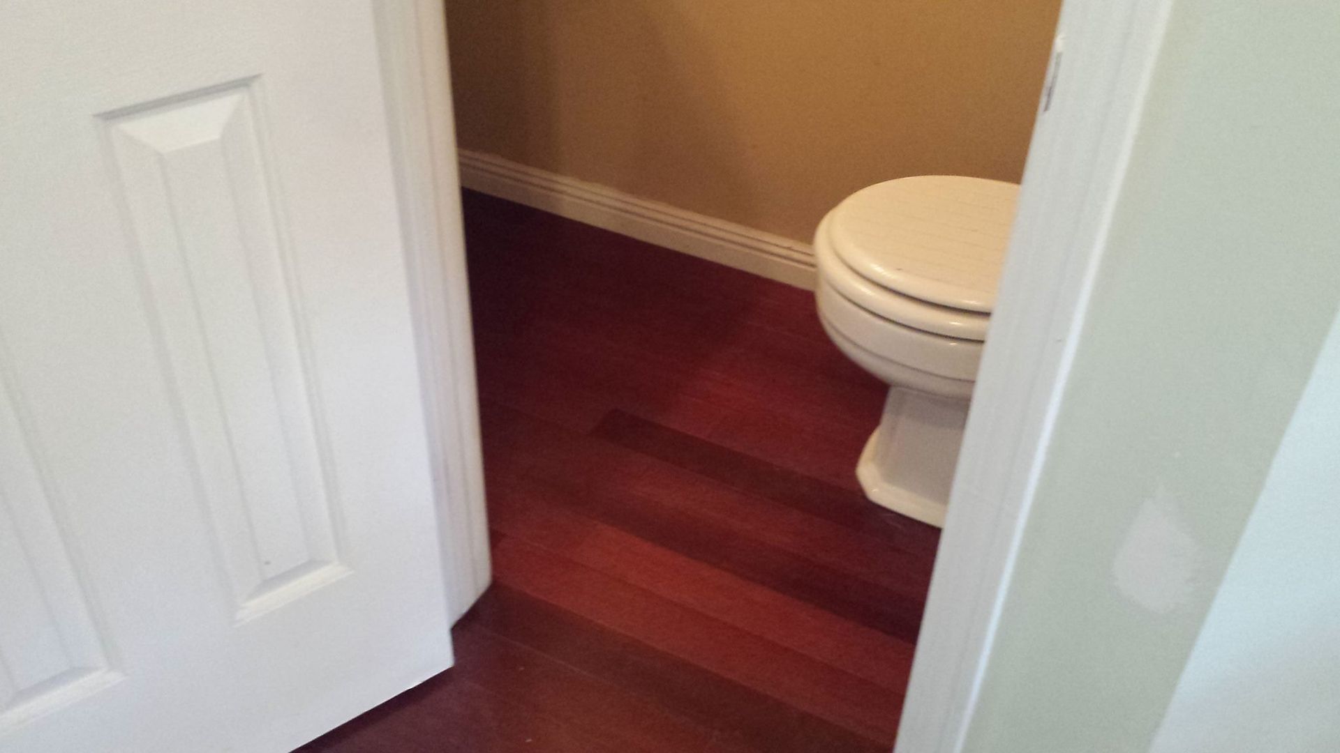 Open doorway reveals a small bathroom with a white toilet on a dark wood floor. Light tan walls.