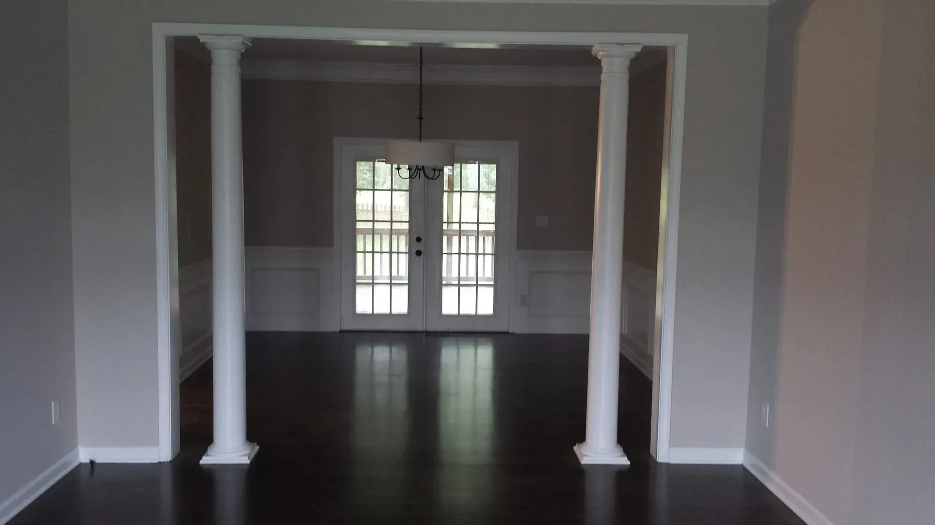 Interior view: dark wood floor, grey walls, white columns framing a doorway to a dining room with white double doors.
