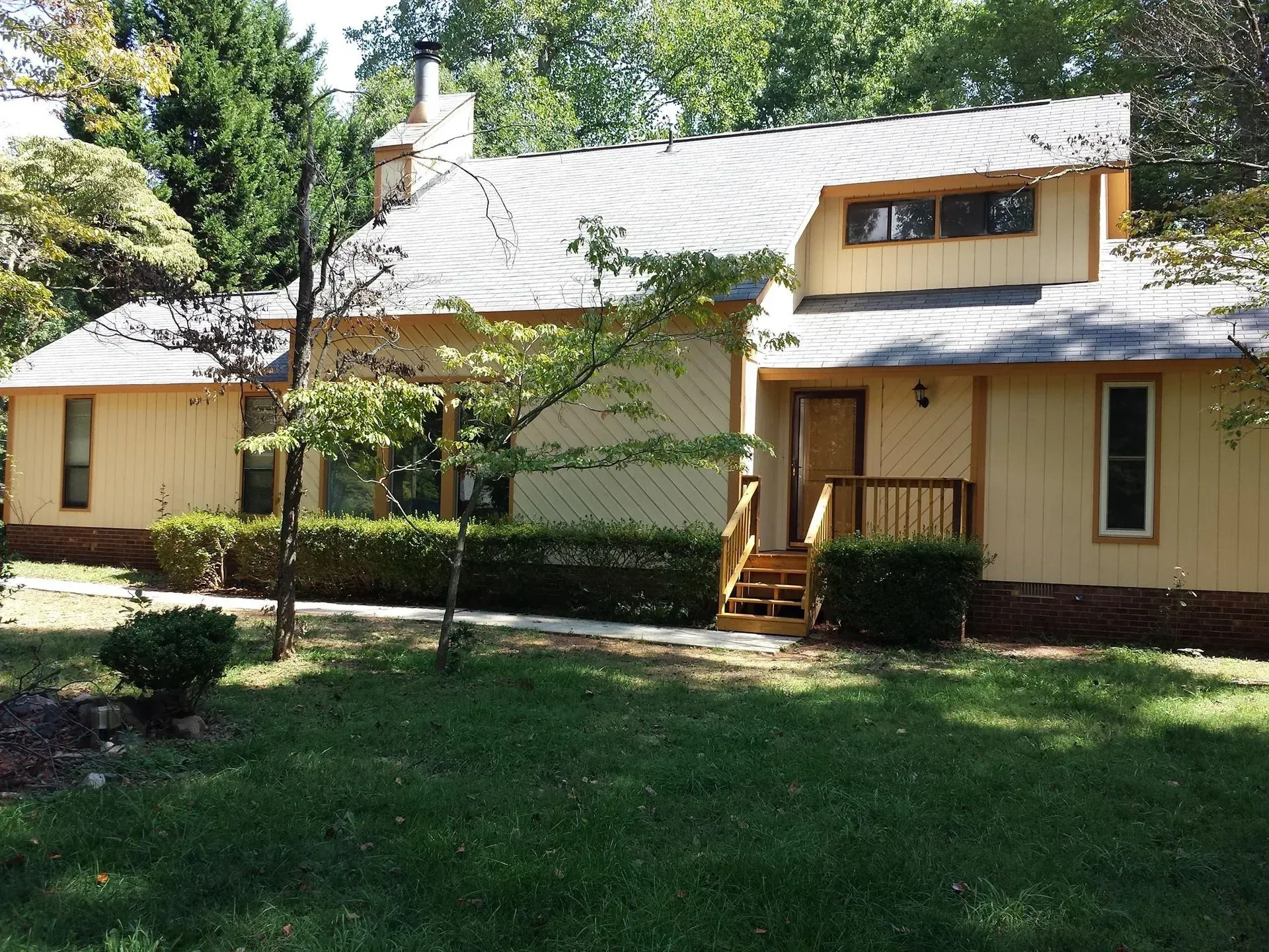 Beige two-story house with brown trim and a gray roof, surrounded by green grass and trees.