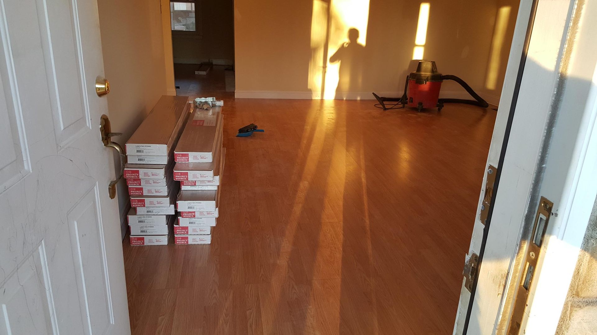 Open doorway into a room with new wood flooring; boxes of flooring in front; person's shadow visible.