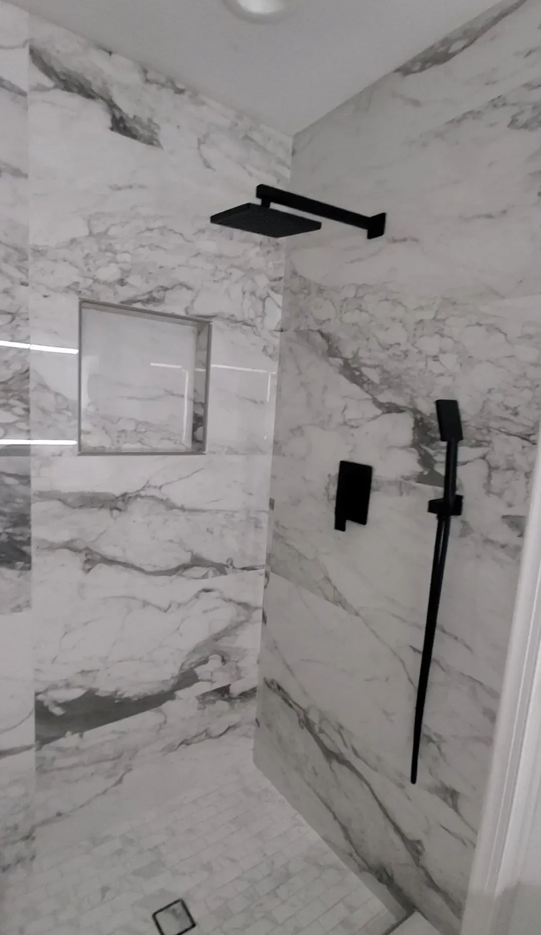Modern shower with marble-look tile, black fixtures, and a recessed shelf.