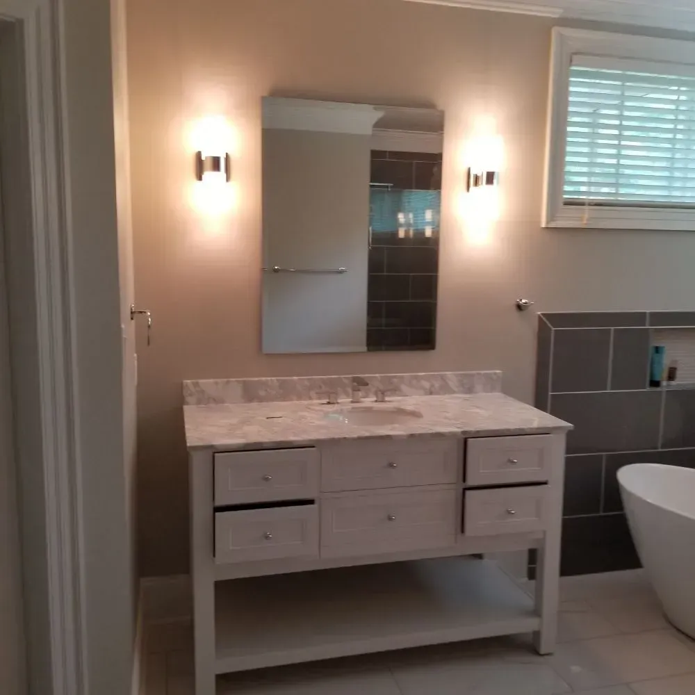 Bathroom with a white vanity, a mirror, and two sconces flanking it. Gray and white tiles are visible.