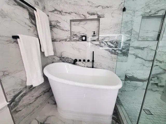 A modern bathroom with a white tub, marble walls, glass shower, and black fixtures.