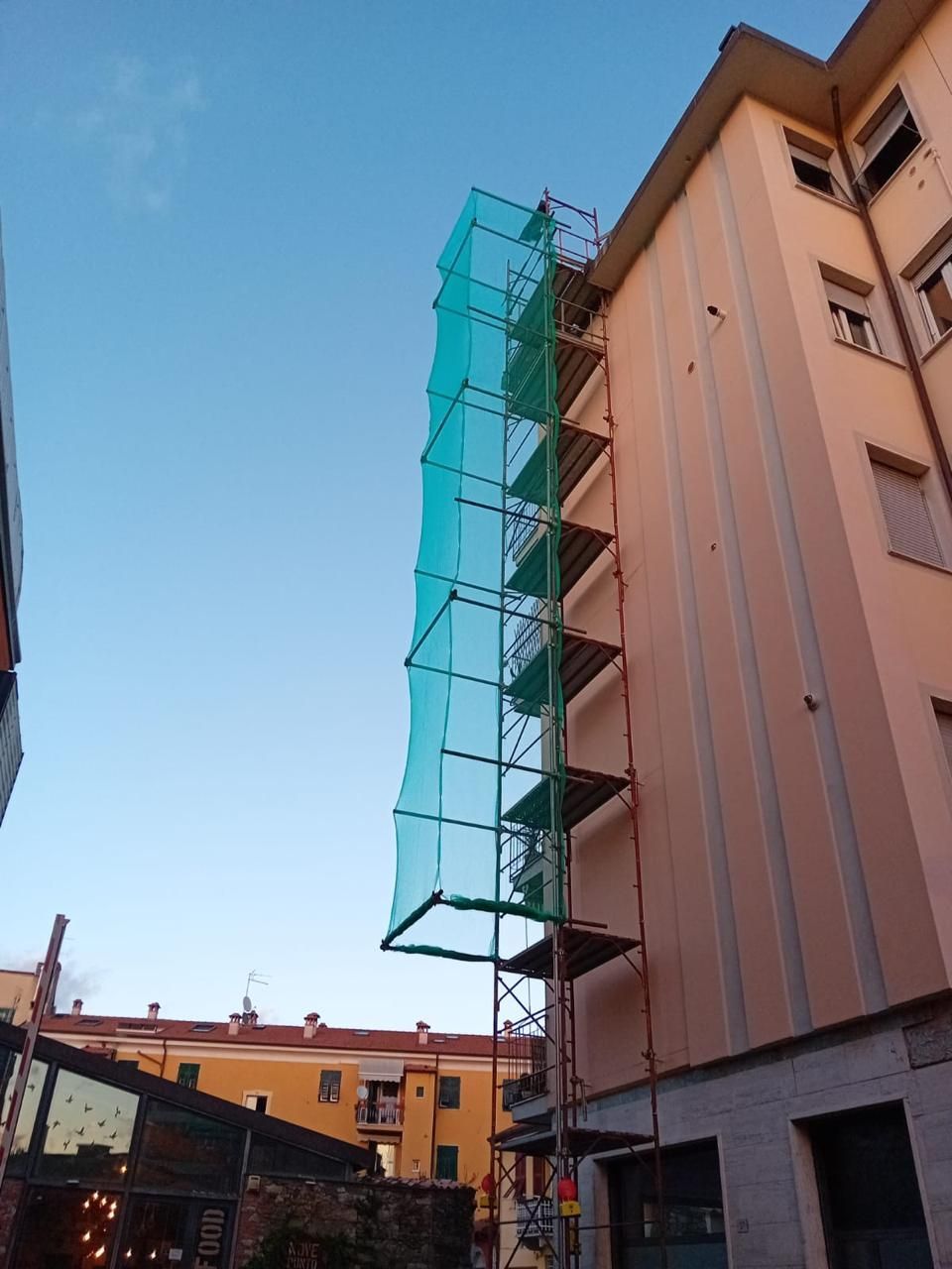 Edificio avvolto da impalcature verdi accanto a un condominio color pesca sotto un cielo azzurro limpido.