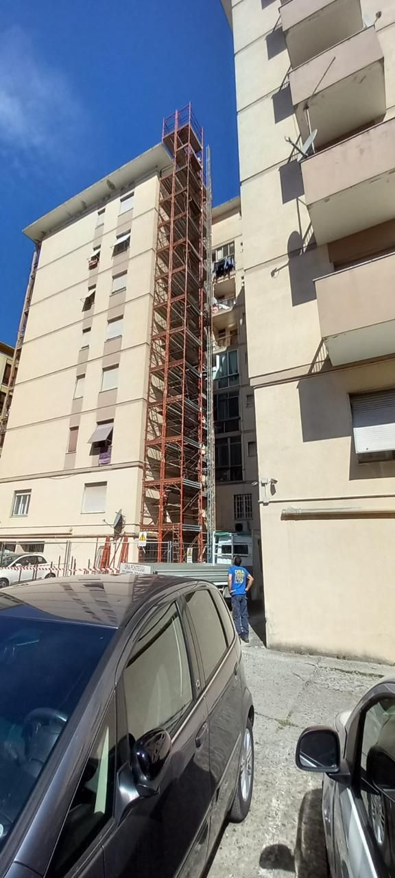 Alto palazzo residenziale con impalcature esterne accanto ad auto parcheggiate in una giornata di sole.