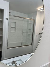 Oval mirror reflecting a white bathroom with a tub/shower, built-in shelves, and a marble countertop.