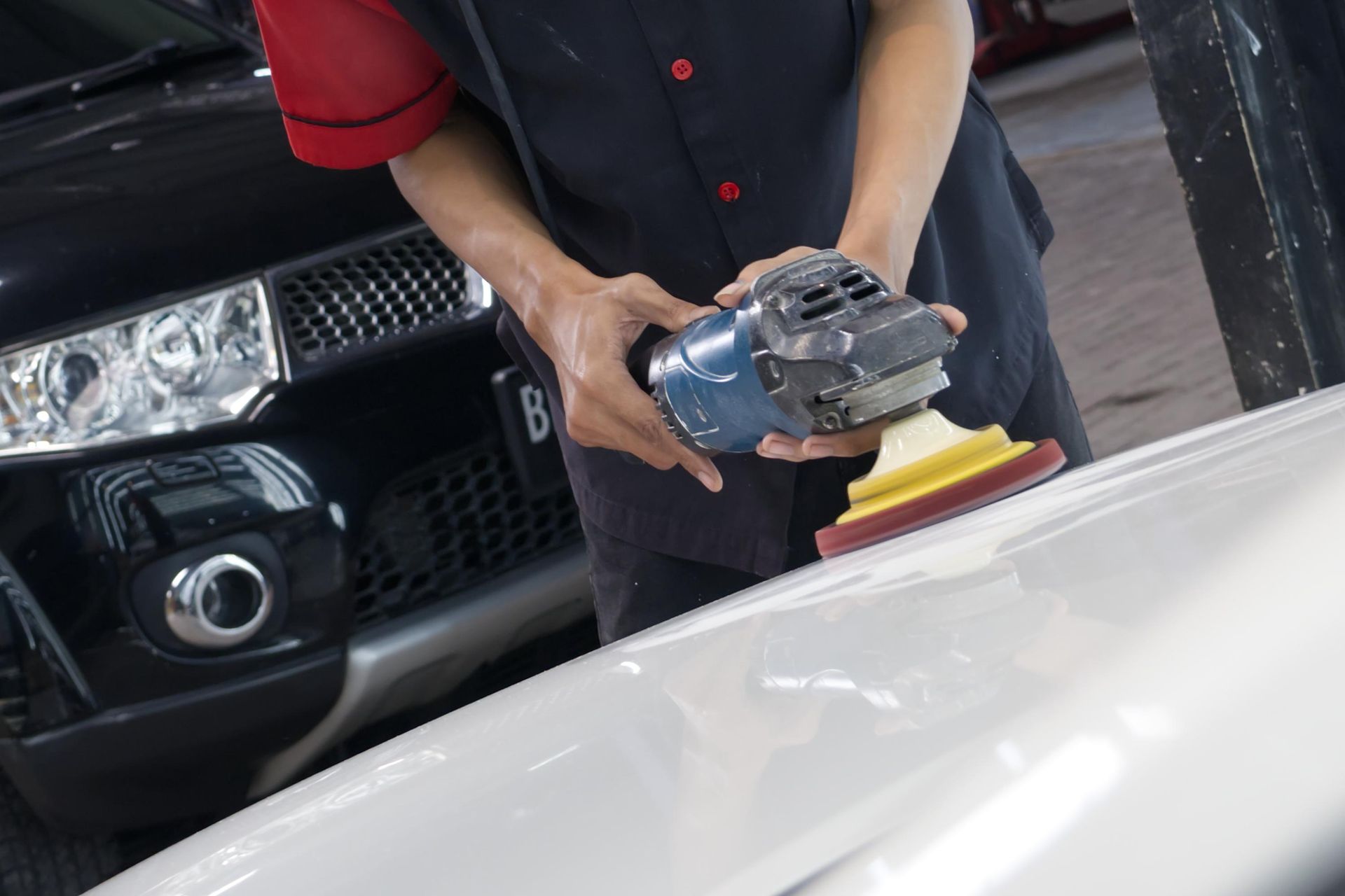 Mechanic polishing a white car hood with a buffer near a black vehicle in a garage