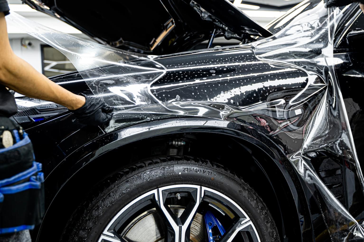 Person applying clear protective film to a black car fender in a workshop