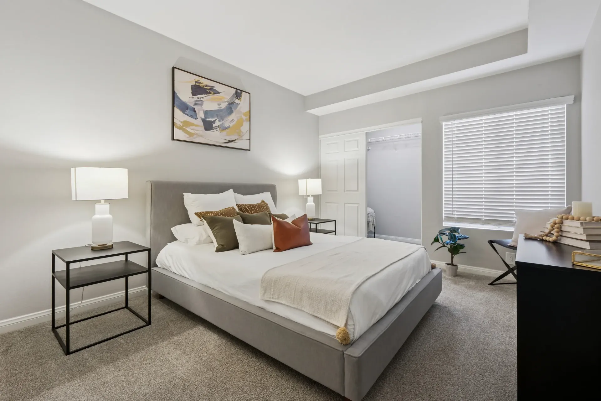 Bedroom in a modern apartment with a gray upholstered bed, two nightstands, and window blinds.