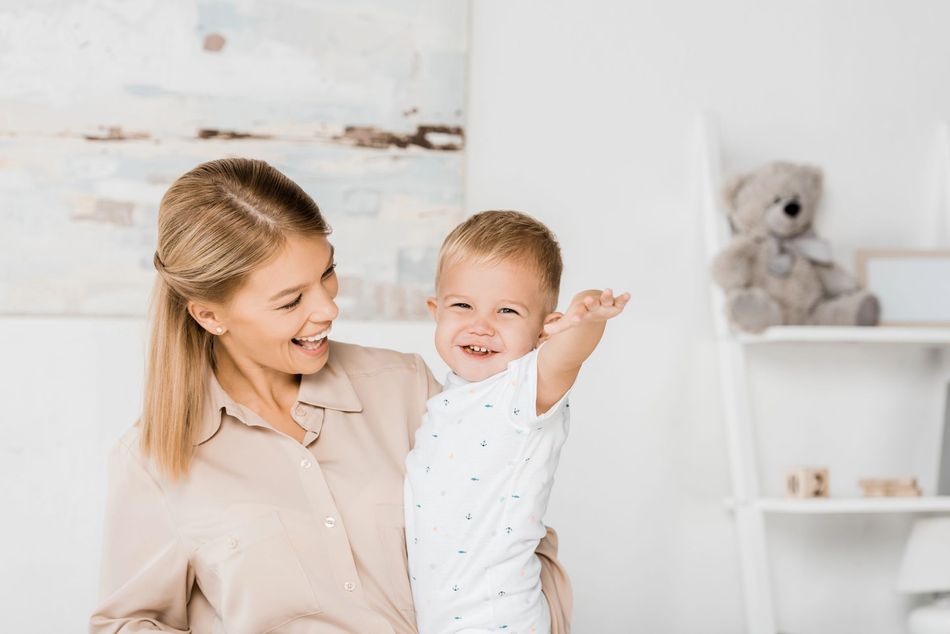 Un adulto sorridente tiene in braccio un bambino felice in una cameretta luminosa, con un orsacchiotto su uno scaffale.