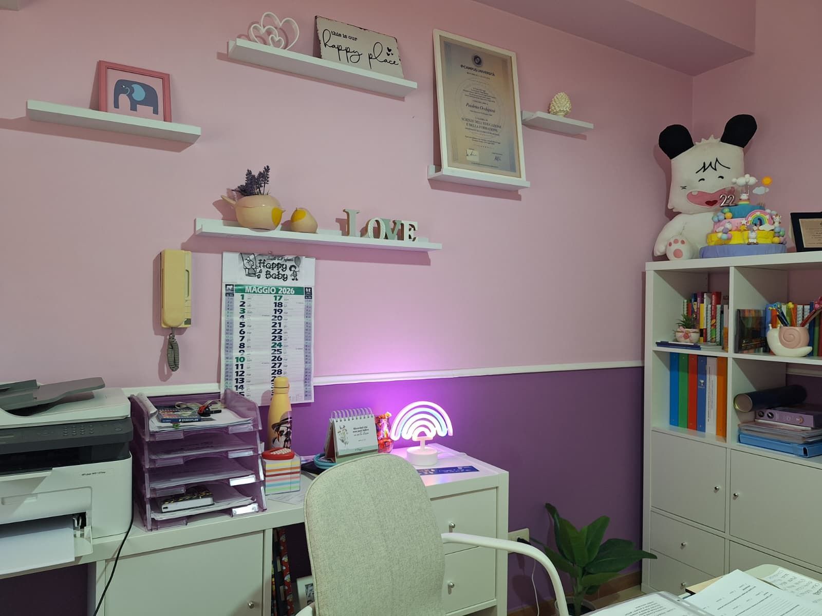 Ufficio domestico rosa con scrivania, sedia bianca, scaffali e un peluche di panda su una libreria bianca.
