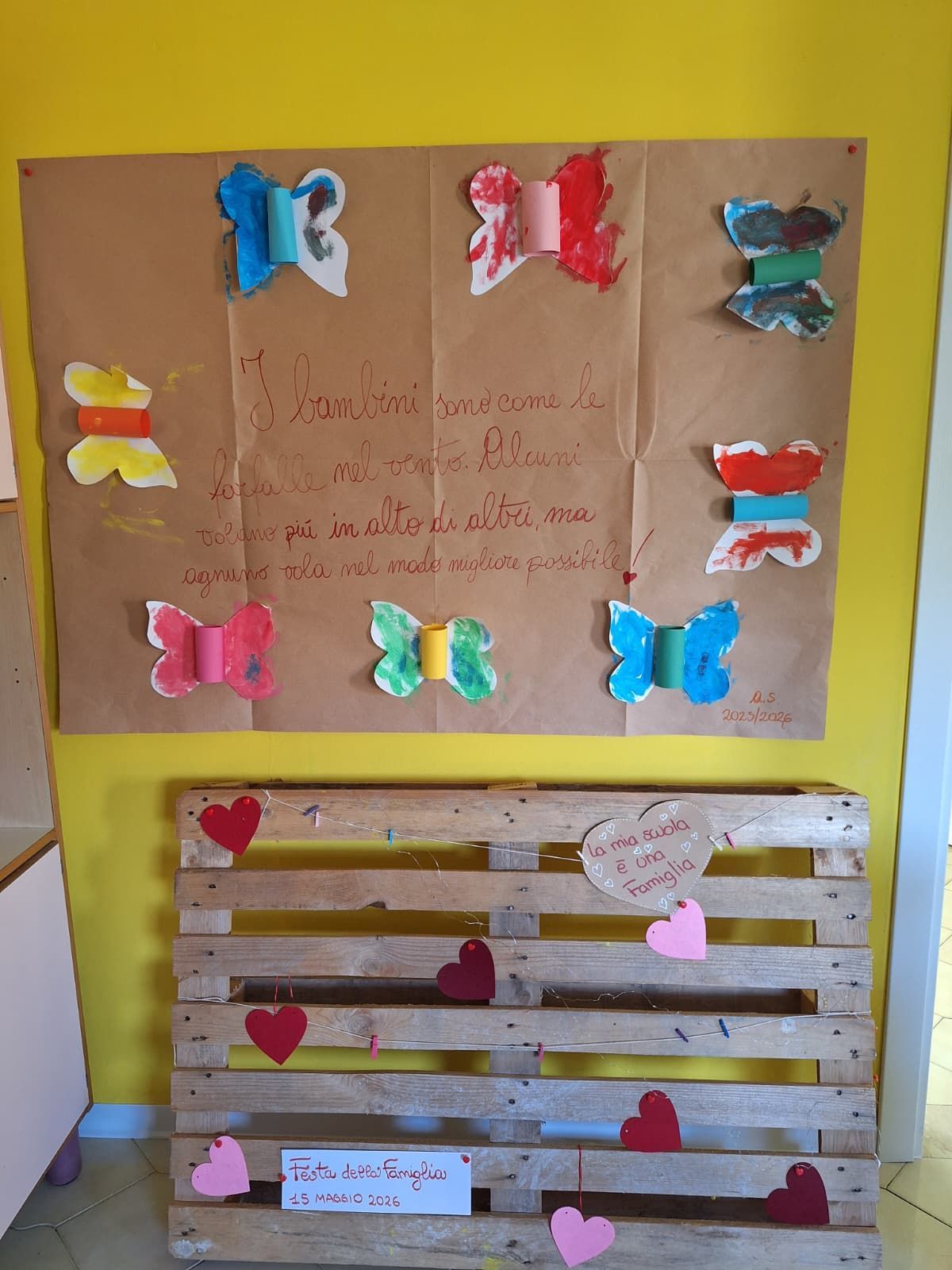 Bacheca decorata con fiocchi e cuori di carta colorata su una parete gialla.