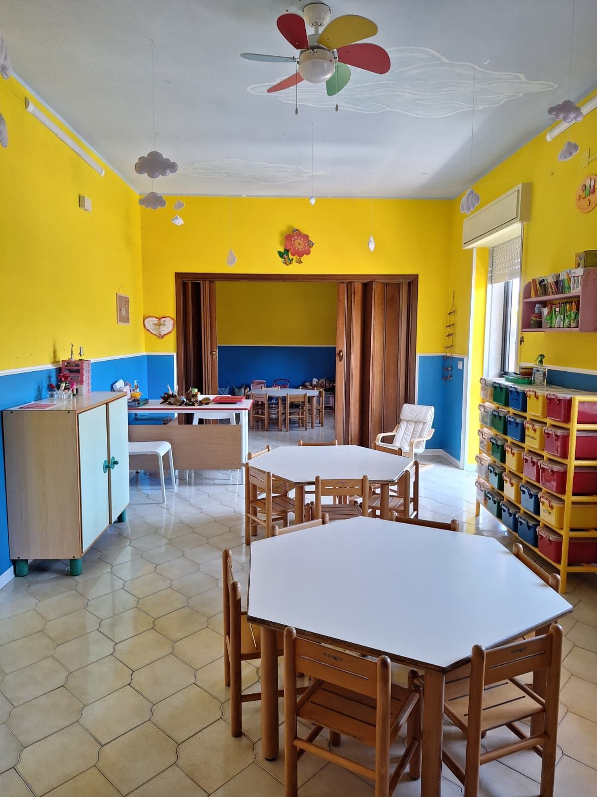 Aula luminosa con pareti gialle e blu, tavoli, sedie di legno e scaffali colorati per riporre gli oggetti.