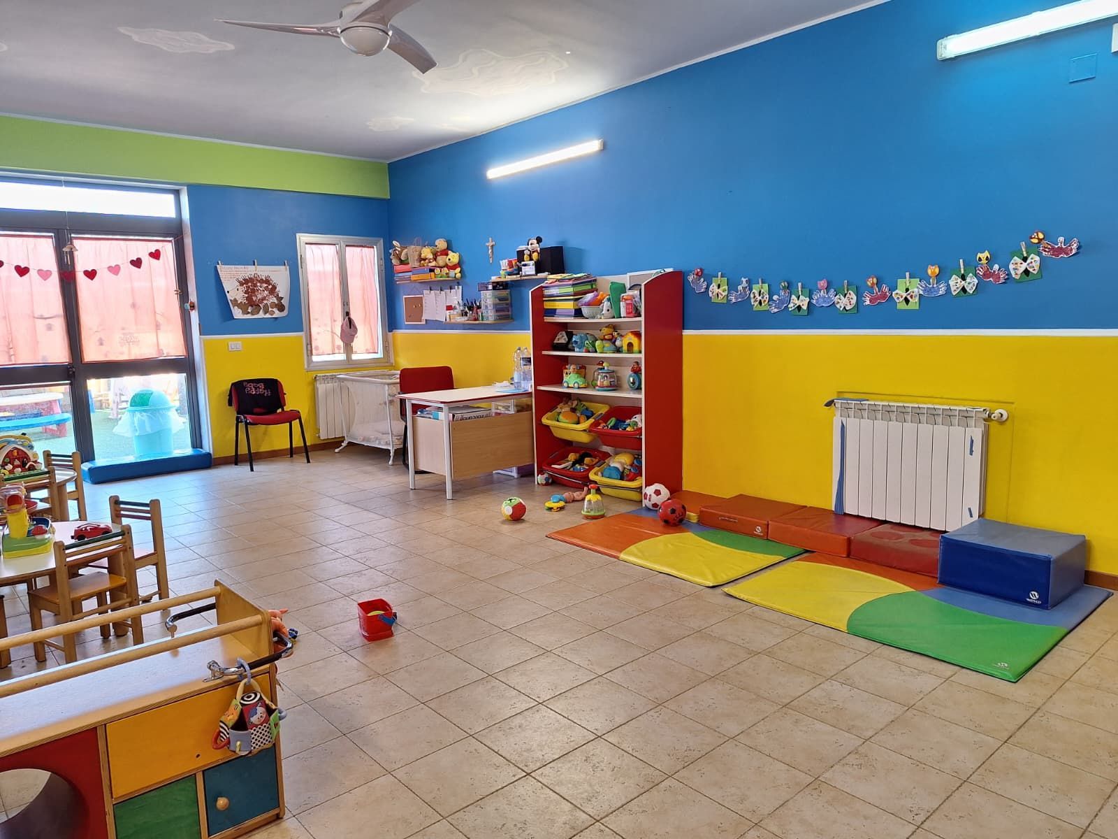 Aula luminosa e colorata dell'asilo nido, con giocattoli, tavoli, sedie, uno scaffale per i giochi e tappeti sul pavimento.