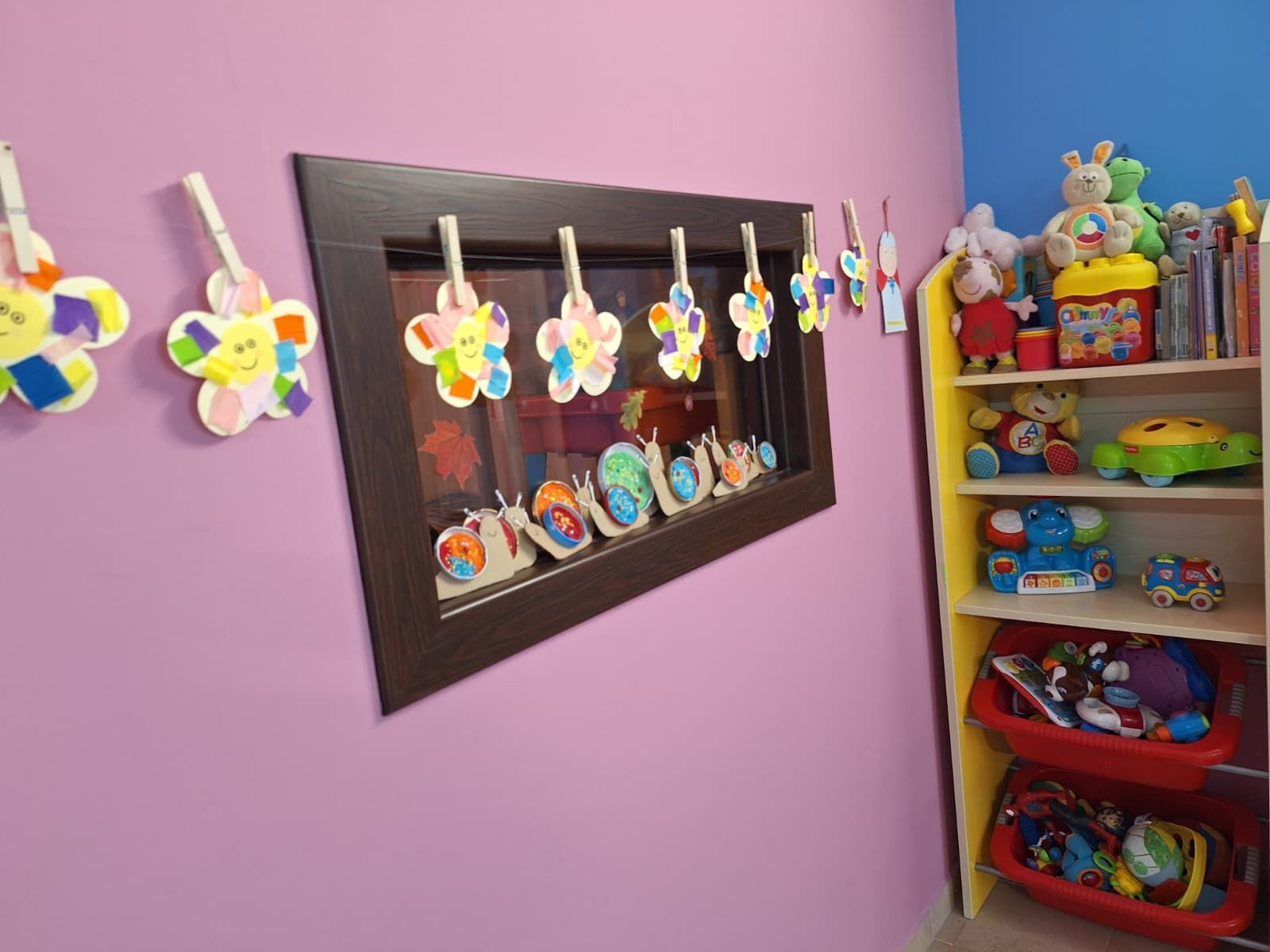 Parete rosa dell'aula con specchio decorato e colorati oggetti artigianali esposti su mensole e ganci.