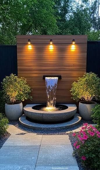 Fountain with tiered basins, water spraying upwards. Green trees, wooden fence and building in the background.