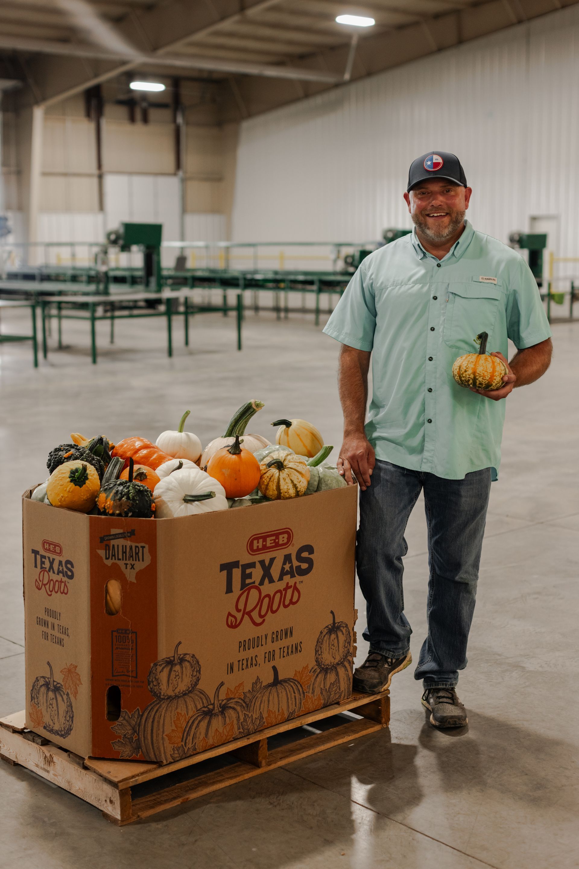 Man standing next to a box of pumpkins in a warehouse. He holds a pumpkin. The box says 
