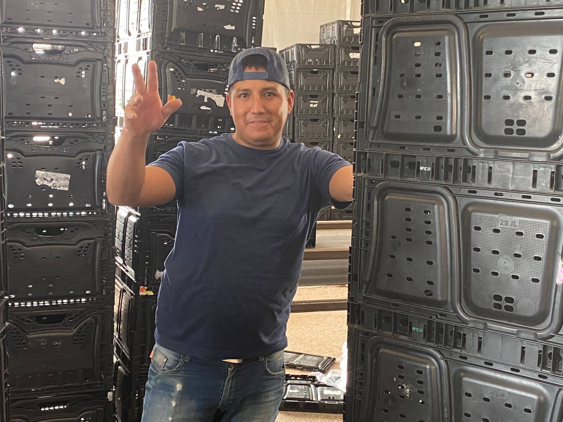 Man in blue shirt and hat, making a peace sign, stands in a warehouse with stacks of black containers.