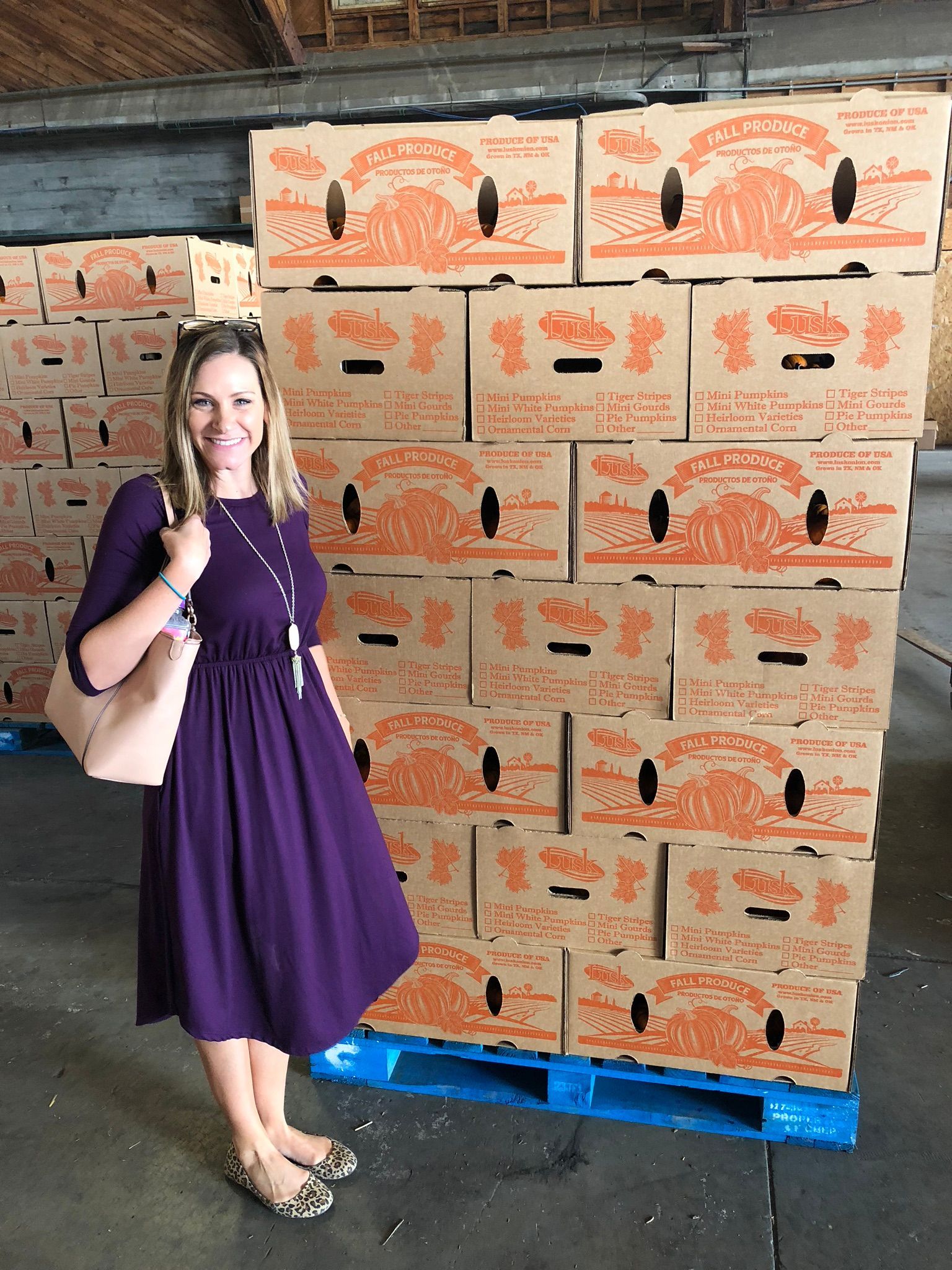 Woman stands next to a tall stack of cardboard boxes inside a warehouse. She wears a purple dress.