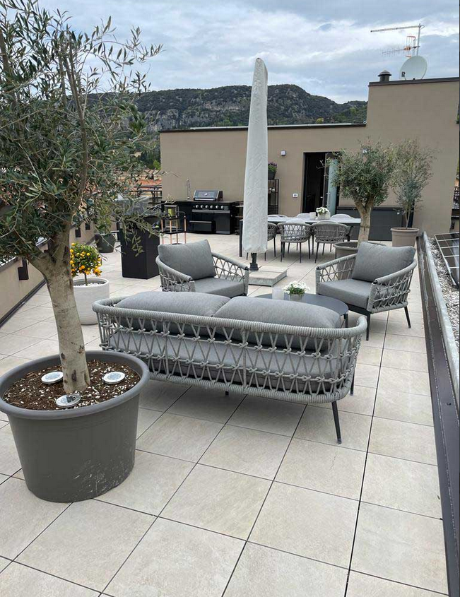 Patio sul tetto con mobili in vimini grigio, ulivi in vaso e vista sulle montagne.