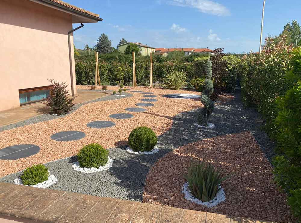 Giardino con ghiaia arancione e grigia, sentiero con gradini in pietra e vegetazione ben curata.