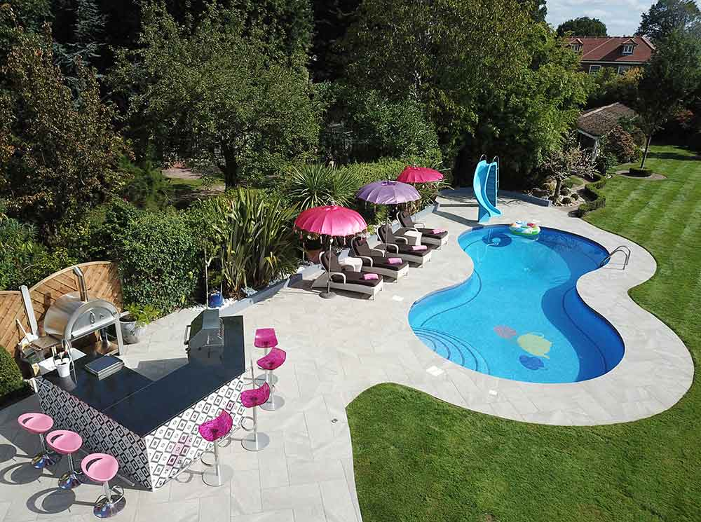 Giardino con piscina, bar, sedie a sdraio e ombrelloni. Prato verde e alberi circondano lo spazio.