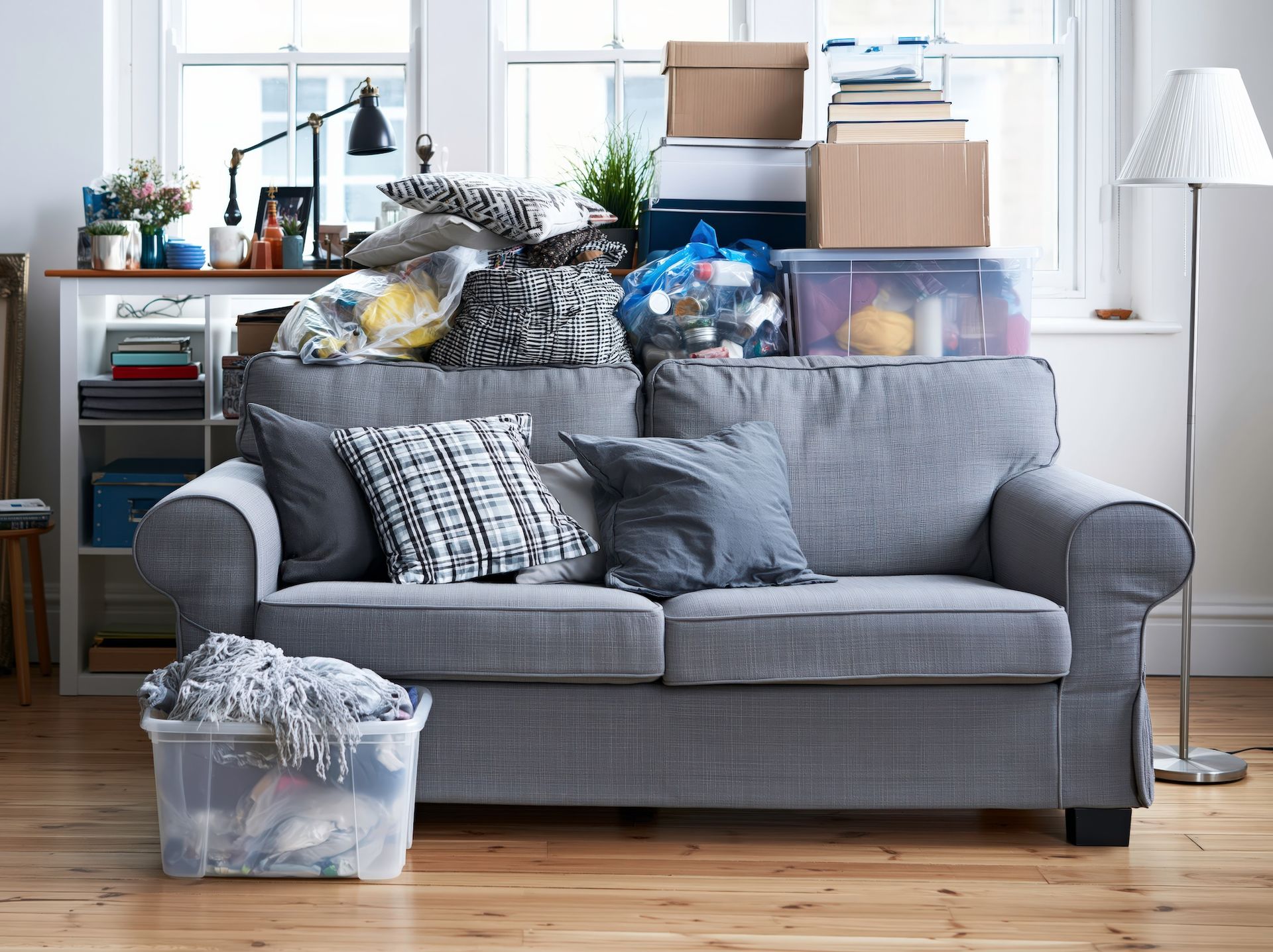 Cluttered gray couch in a messy living room with stacked boxes and household items by a bright window