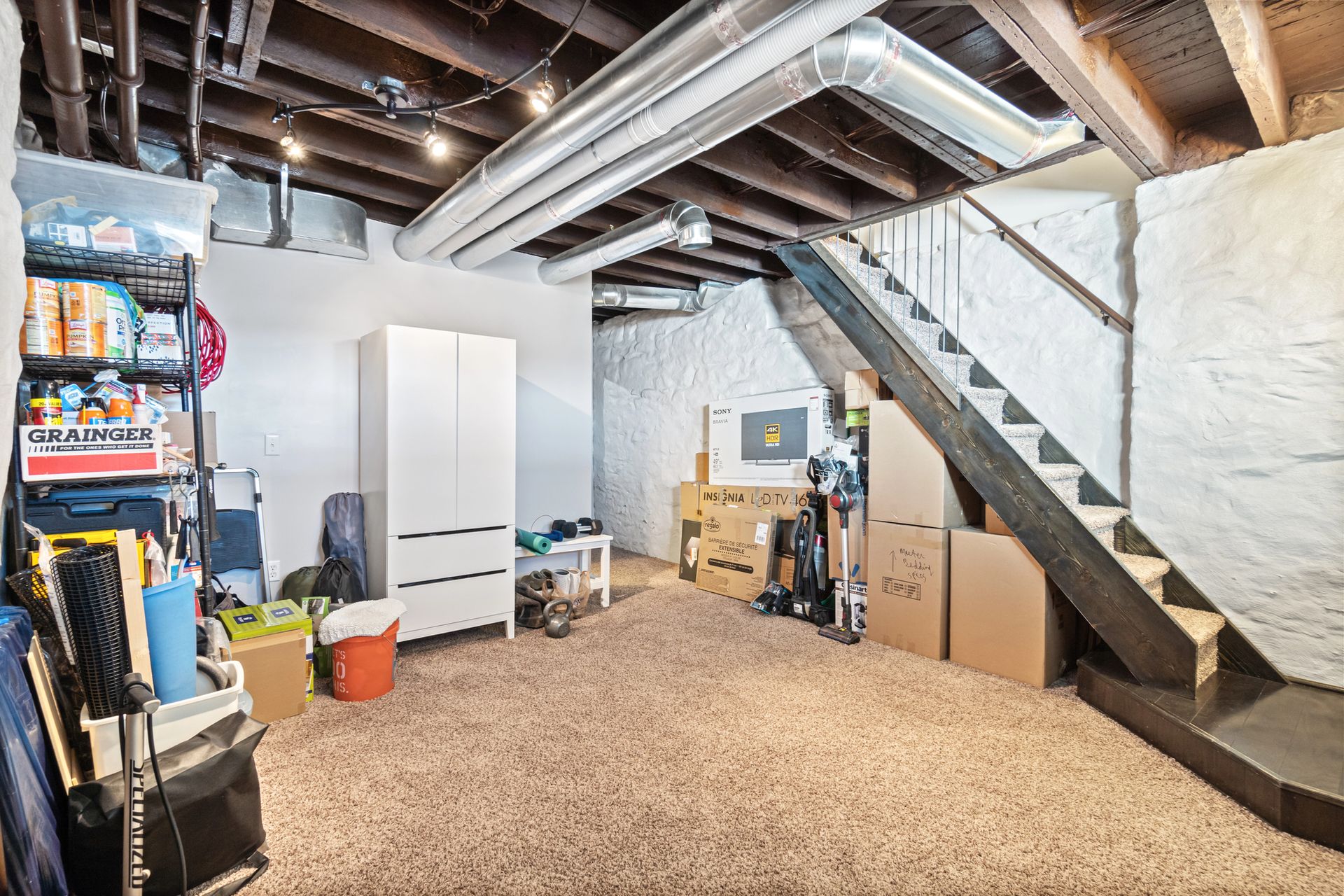 Unfinished basement with exposed beams, storage shelves, stairs, and carpeted floor.
