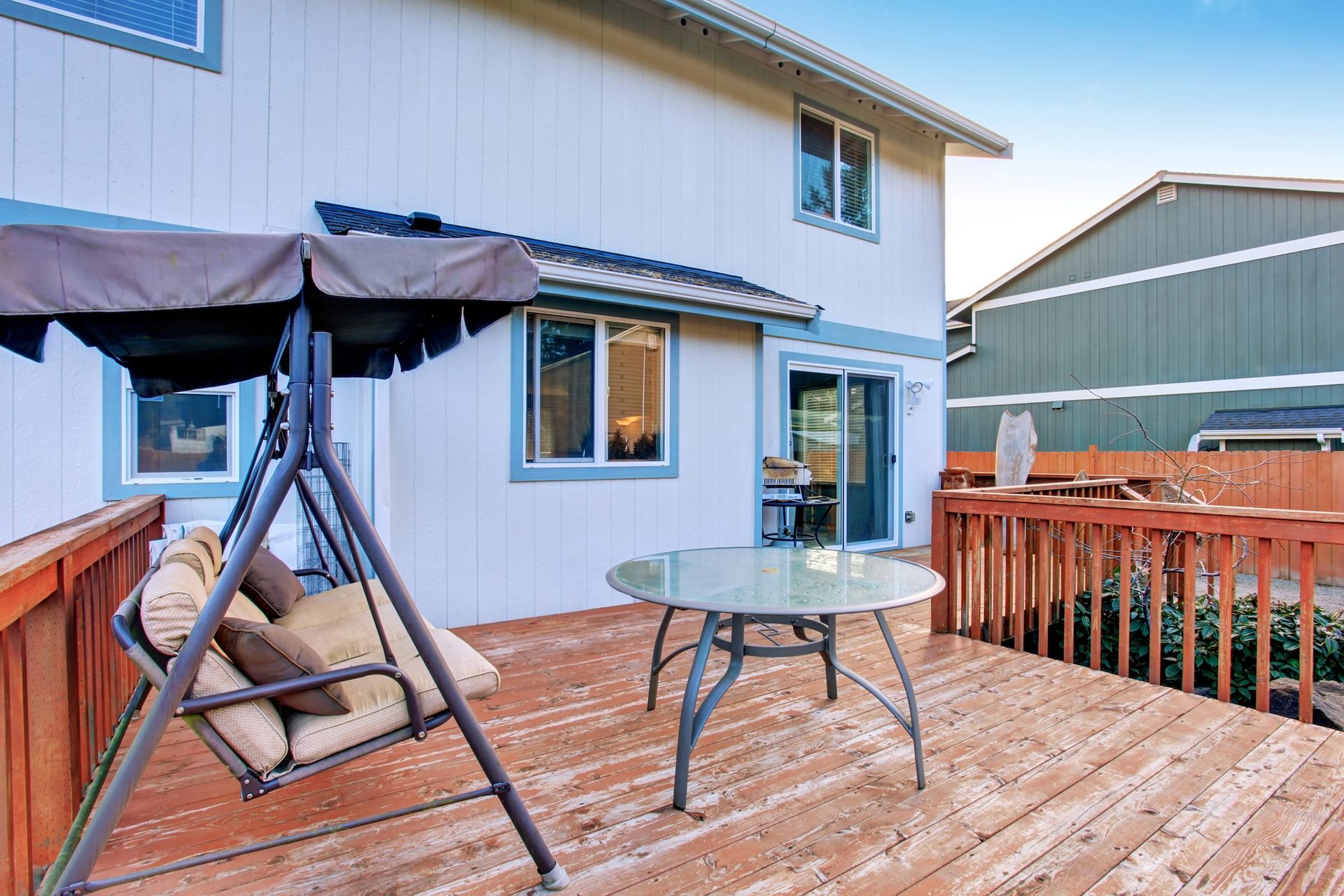 Back deck with swing chair, glass table, and wooden railing beside a white house