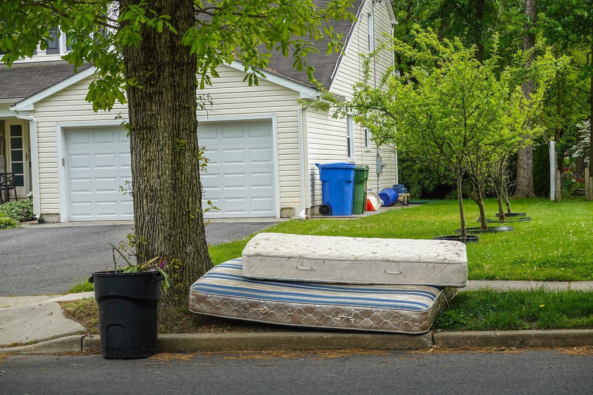 Mattress and box spring left at curb outside a suburban house, beside a tree and trash bin.