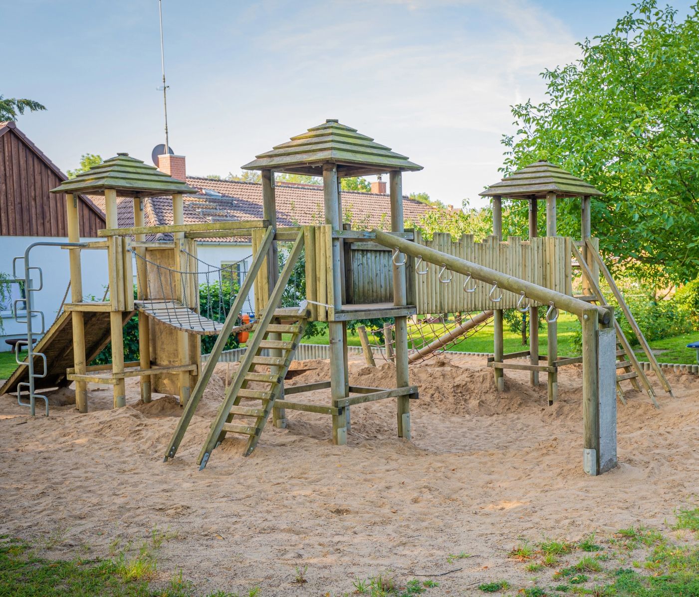 Wooden playground climbing structure with slides and bridges on a sandy surface, surrounded by trees