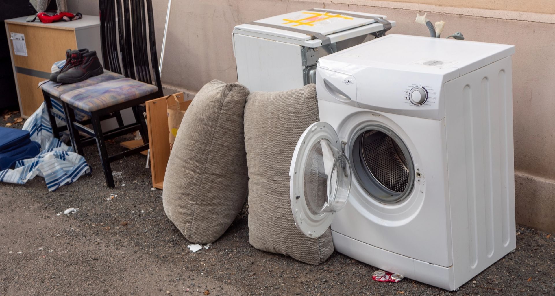 White front-loading washer on carpet next to two beige cushions and a small table in a cluttered room