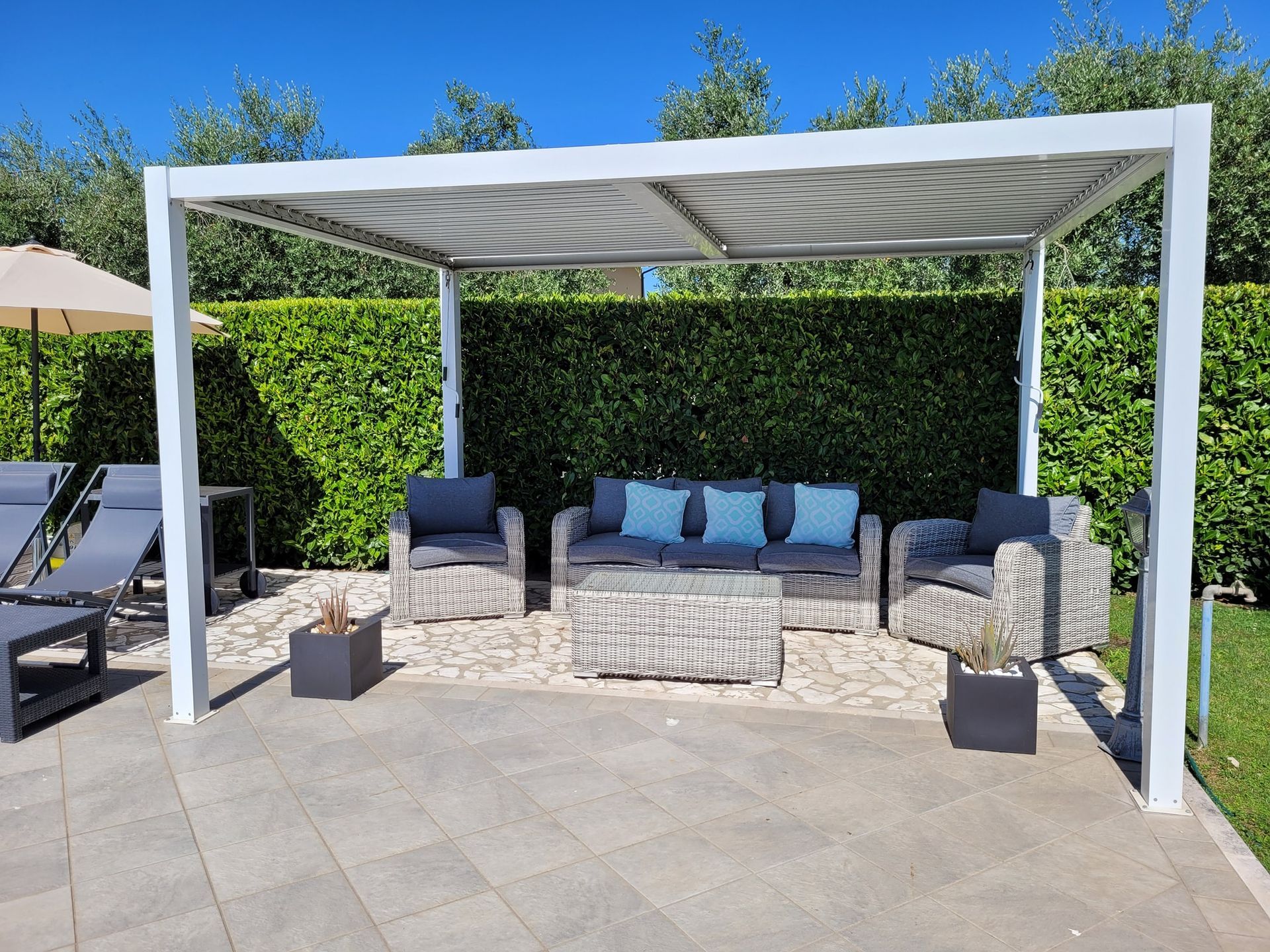 Pergola bianca sopra l'area salotto all'aperto di un patio. Sfondo con siepe verde.