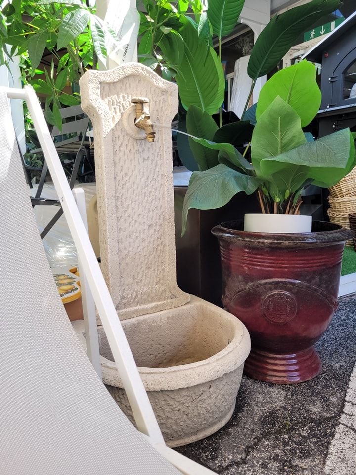 Fontana in pietra con rubinetto in ottone, accanto a una pianta in vaso. In primo piano la struttura di una sedia bianca.