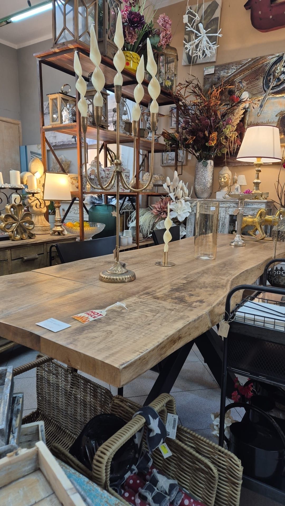 Lungo tavolo di legno in un negozio con candele, fiori e scaffali pieni di decorazioni.