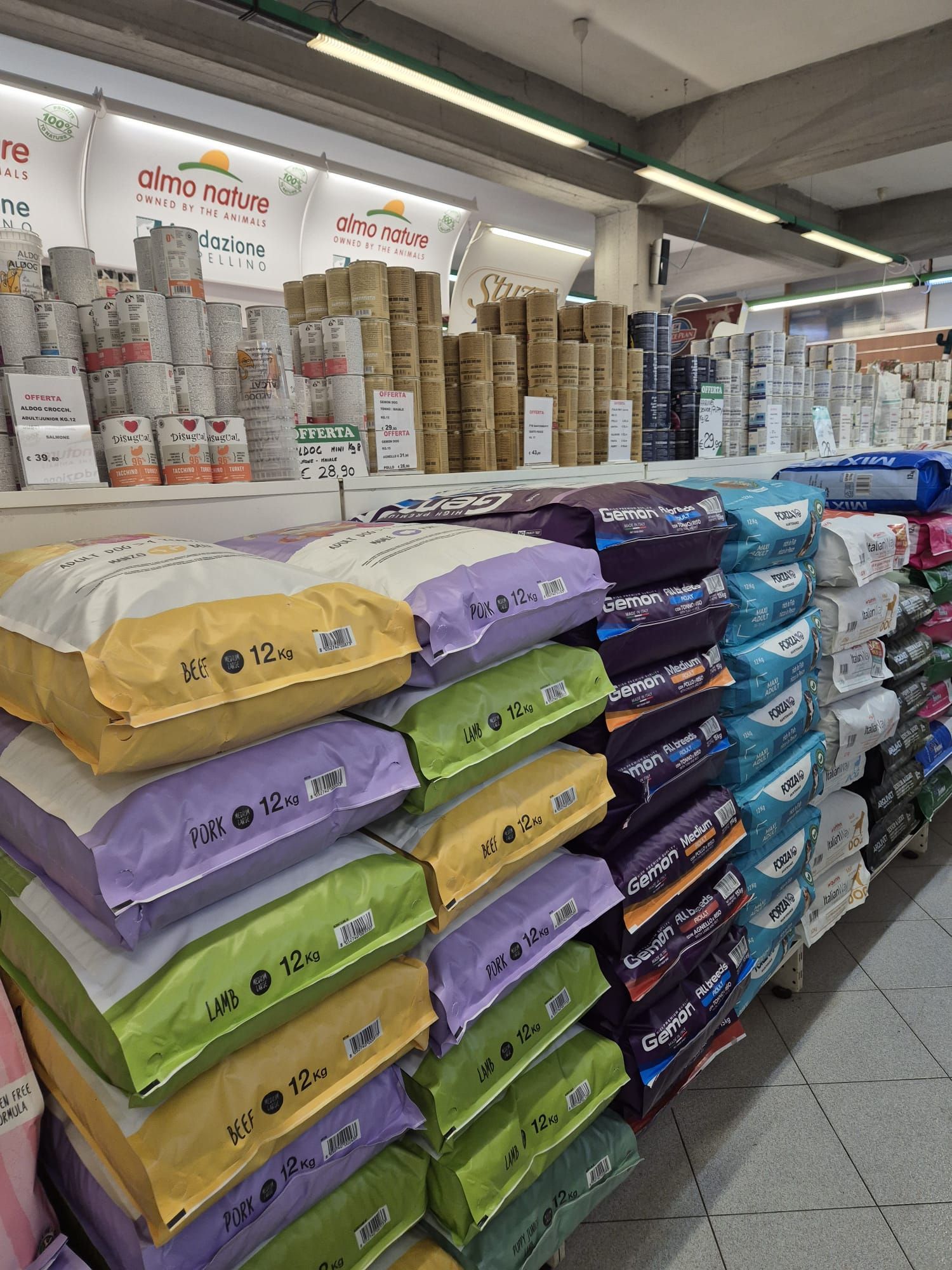 Sacchi di cibo per animali domestici accatastati sugli scaffali di un negozio, con vari colori e marche visibili.