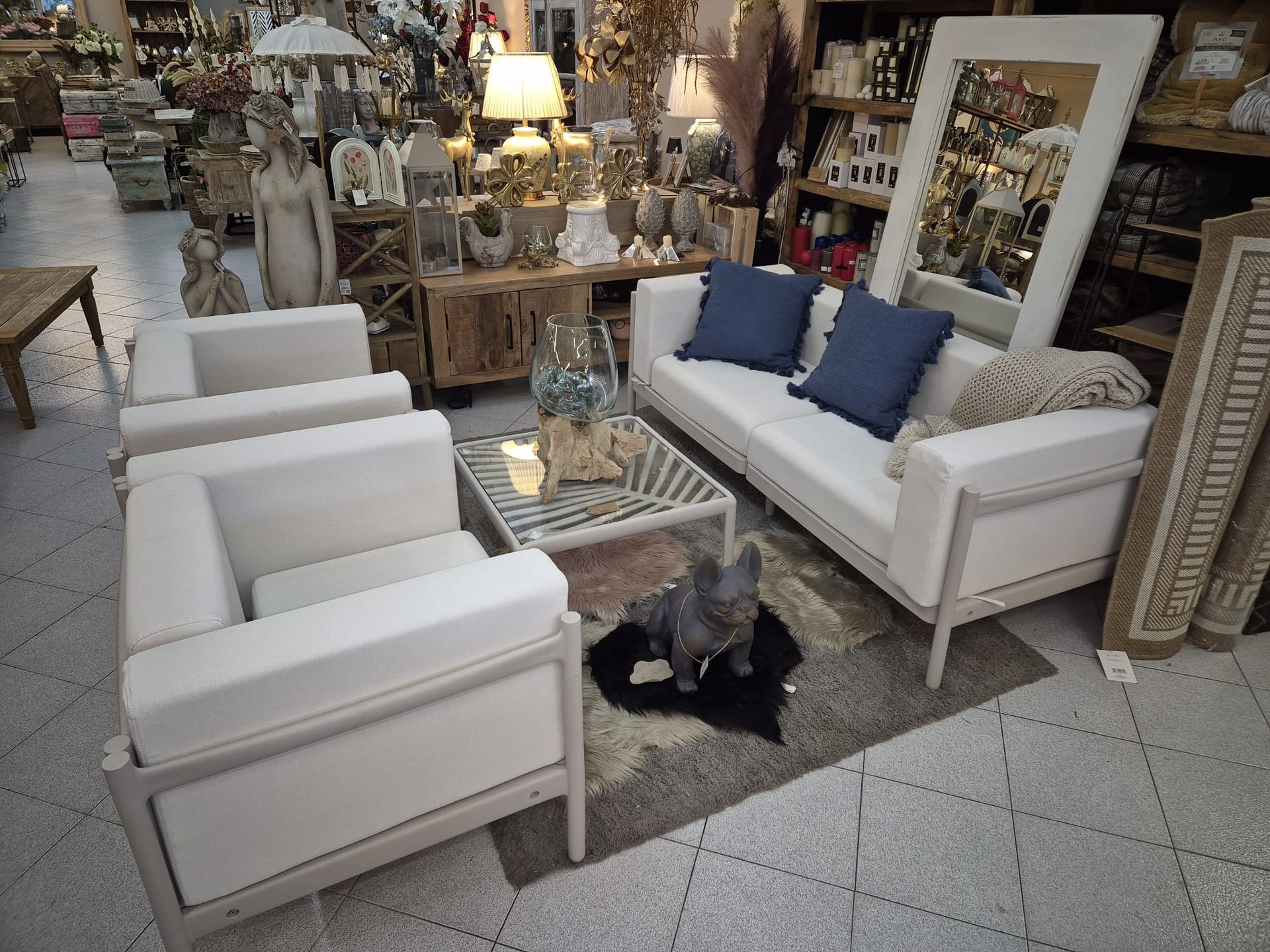Soggiorno con divani e poltrone bianchi, cuscini blu, un cagnolino su un tappeto e vari oggetti decorativi in un negozio.