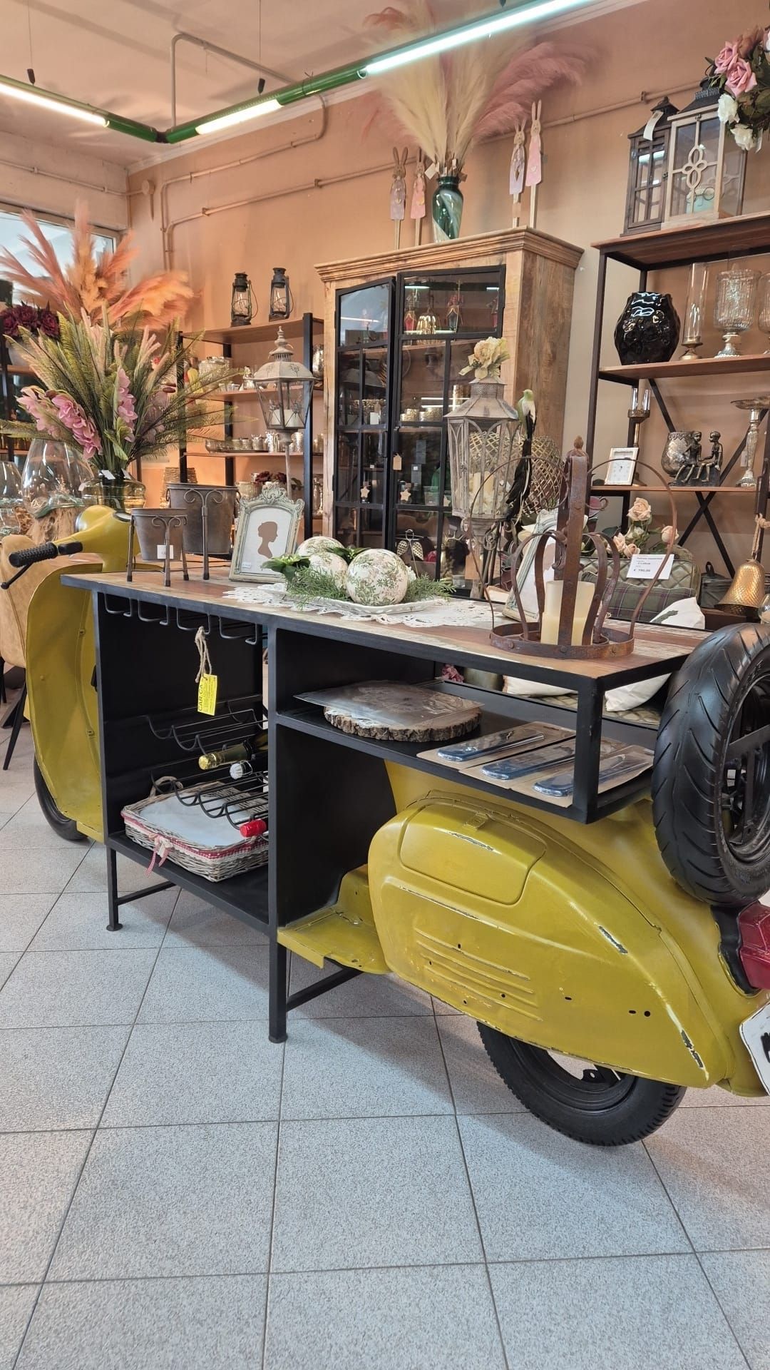 Scooter giallo trasformato in un bancone espositivo per negozio con scaffali, vari articoli e uno sfondo marrone chiaro.