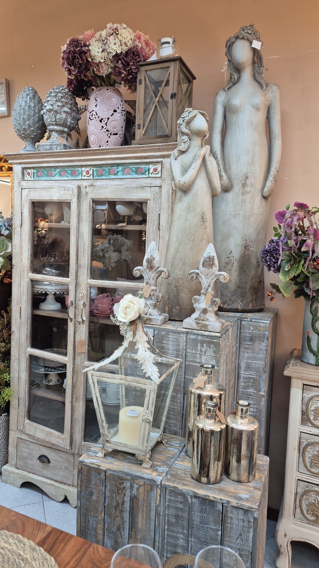 Esposizione di oggetti decorativi, tra cui un mobile, statue, lanterne e fiori, in un ambiente da negozio.