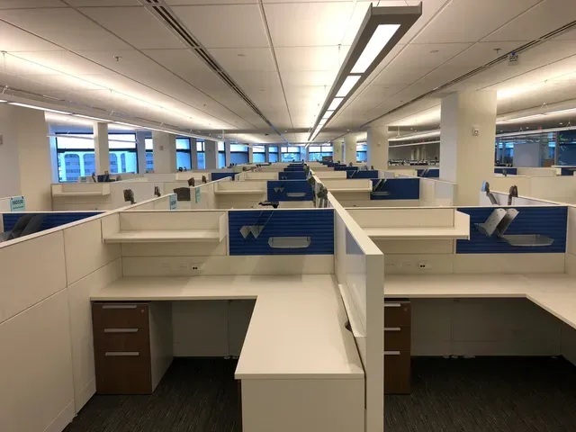 Empty office space with rows of white cubicles featuring blue accents and wooden drawer units under fluorescent lights.
