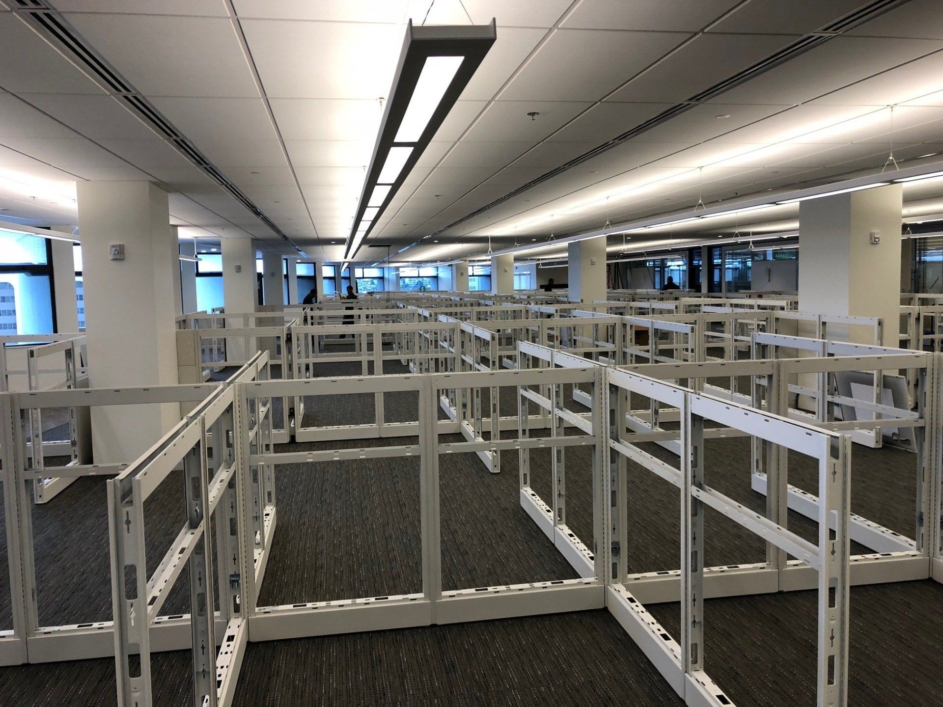 An open-plan office space featuring rows of white cubicle frames set against dark grey carpeting under bright lighting.