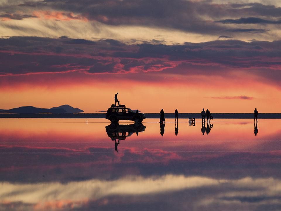 Silhouetted car and people on a salt lake at sunset, with vivid pink and orange sky reflected on water