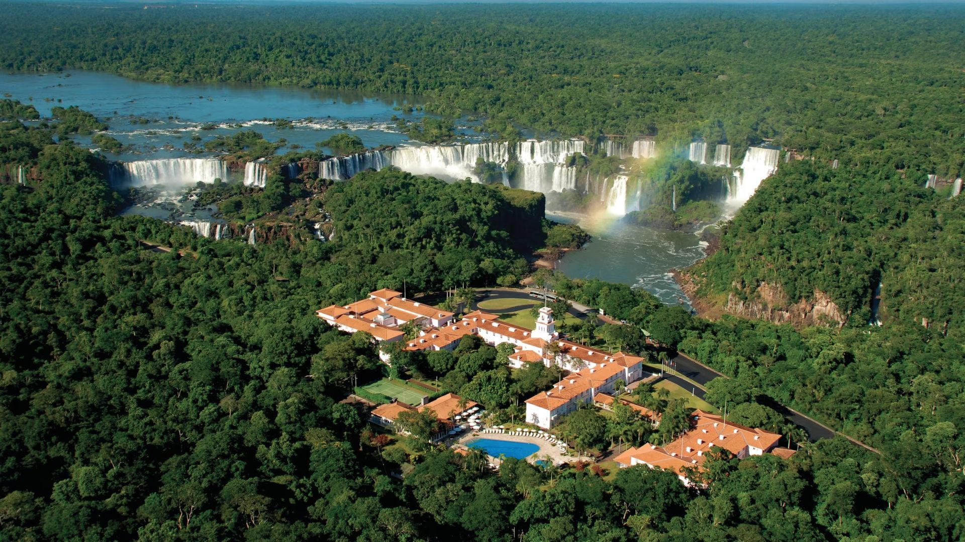Aerial view of a lush rainforest resort near wide waterfalls and river cliffs.