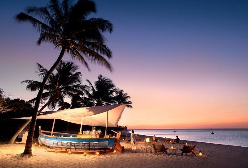 Beach at sunset with palm trees, sailboats on the shore, and people relaxing by the water.