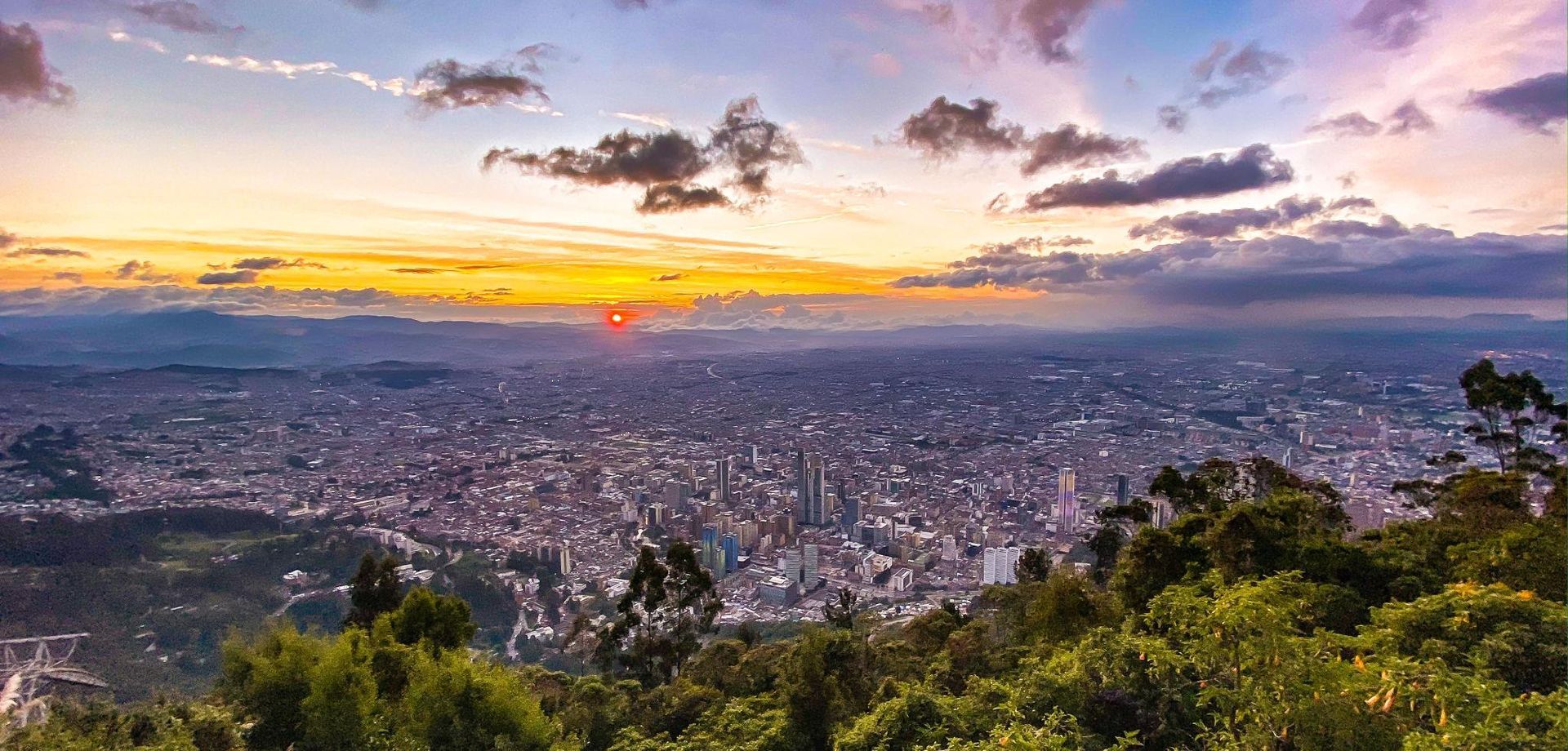 Sunset over the sprawling Bogota cityscape with glowing sky and silhouetted trees in the foreground