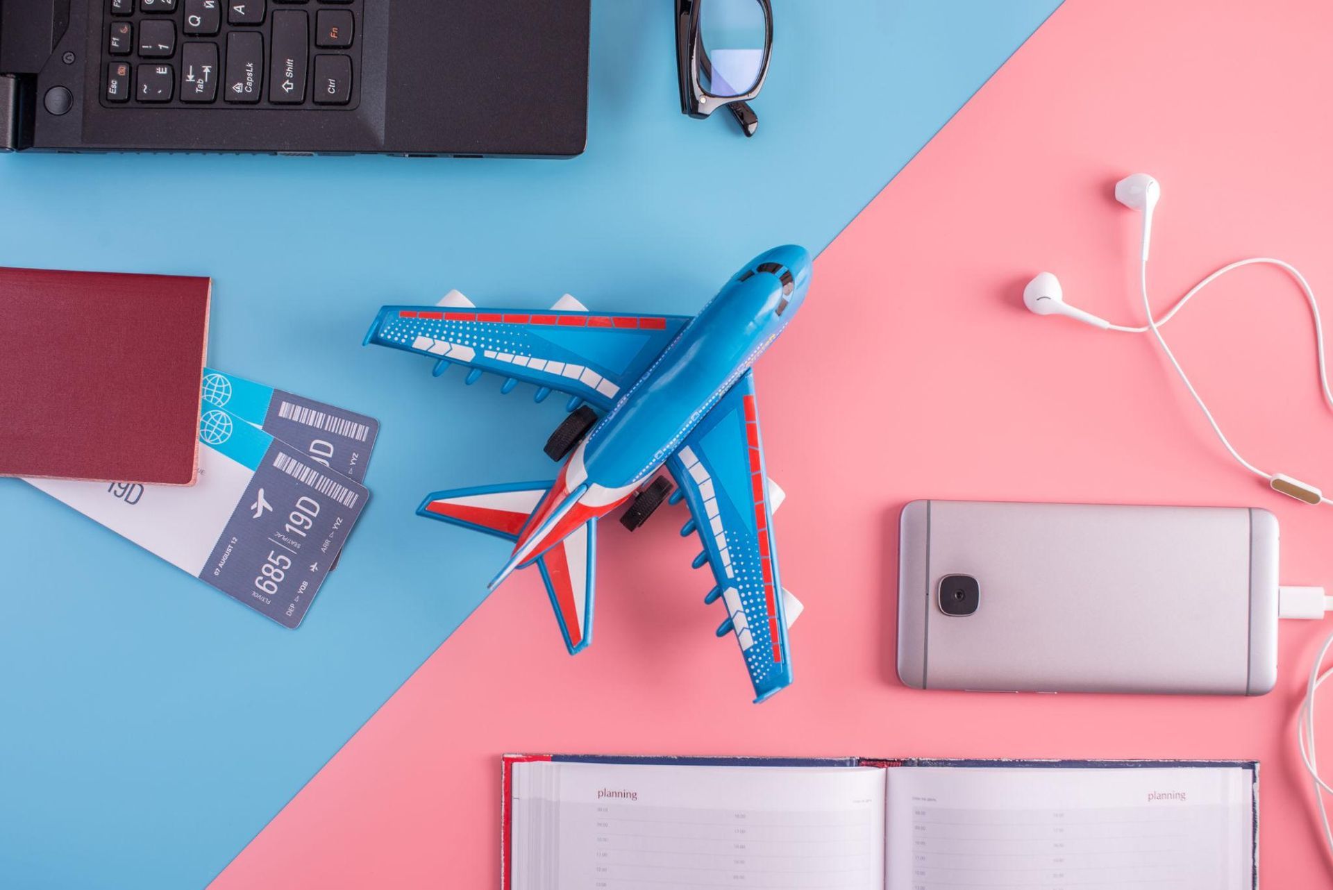 Top-down travel workspace with blue toy airplane, passport, smartphone, notebook, earbuds, and keyboard on pink and blue desk
